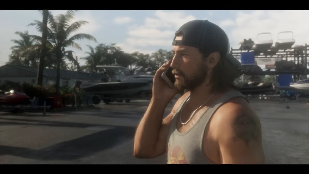 Un homme à casquette retournée et débardeur gris, téléphone à l'oreille, devant des bateaux posés sur des racks.